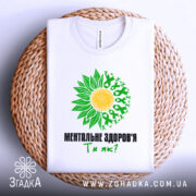 Футболка ментальне здоров`я білого кольору, зображена зверху на плоскій поверхні, оточена плетеним кошиком. Принт з жовтим сонцем та зеленими листками, текст чорного кольору: 'МЕНТАЛЬНЕ ЗДОРОВ'Я Ти як?'.