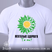 Футболка ментальне здоров`я білого кольору, зображена зверху, на плоскій поверхні. Принт з жовтим сонцем та зеленими листками, текст чорного кольору: 'МЕНТАЛЬНЕ ЗДОРОВ'Я Ти як?'. На футболці підвішена етикетка.