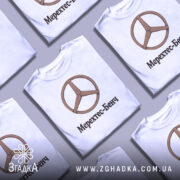 Футболка Мерехтес-Бенч стиль, біла, складена на столі, фон світло-сірий, кілька футболок з дерев'яним логотипом і написом 'Мерехтес-Бенч' розташовані поруч.