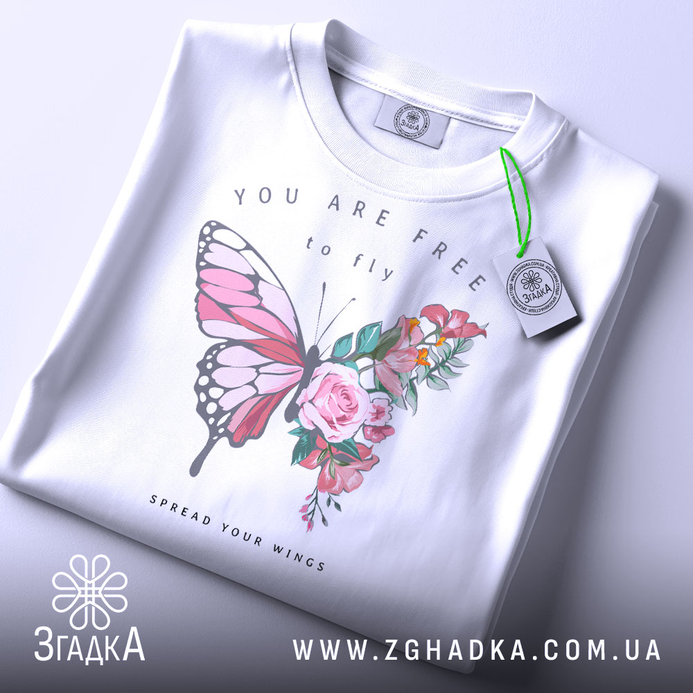 Футболка метелик квіти, біла, складена на столі, фон світло-сірий, принт з метеликом та квітами, текст "YOU ARE FREE to fly" та "SPREAD YOUR WINGS", на футболці є етикетка з зеленим шнурком.