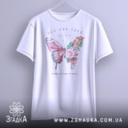 Футболка метелик квіти, біла, підвішена на вішалці, фон світло-сірий, принт з метеликом та квітами, текст "YOU ARE FREE to fly" та "SPREAD YOUR WINGS" під принтом.