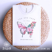 Футболка метелик квіти, біла, складена на плетеному кошику, фон світло-сірий, принт з метеликом та квітами, текст "YOU ARE FREE to fly" та "SPREAD YOUR WINGS", видно етикетку з написом.