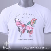 Футболка метелик квіти, біла, складена на столі, фон світло-сірий, принт з метеликом та квітами, текст "YOU ARE FREE to fly" та "SPREAD YOUR WINGS", на футболці є етикетка з зеленим шнурком.