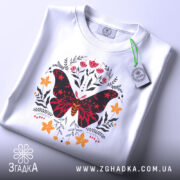 Футболка метелик квіти, біла, зображена зверху, фон світло-сірий, принт з метеликом червоного та чорного кольорів, оточений квітами жовтого та червоного відтінків, на футболці є етикетка з зеленим шнурком.