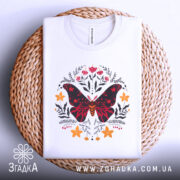 Футболка метелик квіти, біла, зображена зверху на плетеній кошику, фон світло-сірий, принт з метеликом червоного та чорного кольорів, оточений квітами жовтого та червоного відтінків.