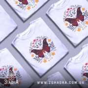 Футболка метелик квіти, біла, зображена на манекені, фон світло-сірий, принт з метеликом червоного та чорного кольорів, оточений квітами жовтого та червоного відтінків.