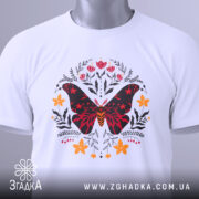 Футболка метелик квіти, біла, зображена зверху, фон світло-сірий, принт з метеликом червоного та чорного кольорів, оточений квітами жовтого та червоного відтінків, на футболці є етикетка з зеленим шнурком.