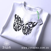 Футболка метелик вітання, біла, складена на поверхні, фон світло-сірий, принт чорного кольору з написом 'HAPPY BIRTHDAY', на футболці є етикетка з зеленим шнурком.