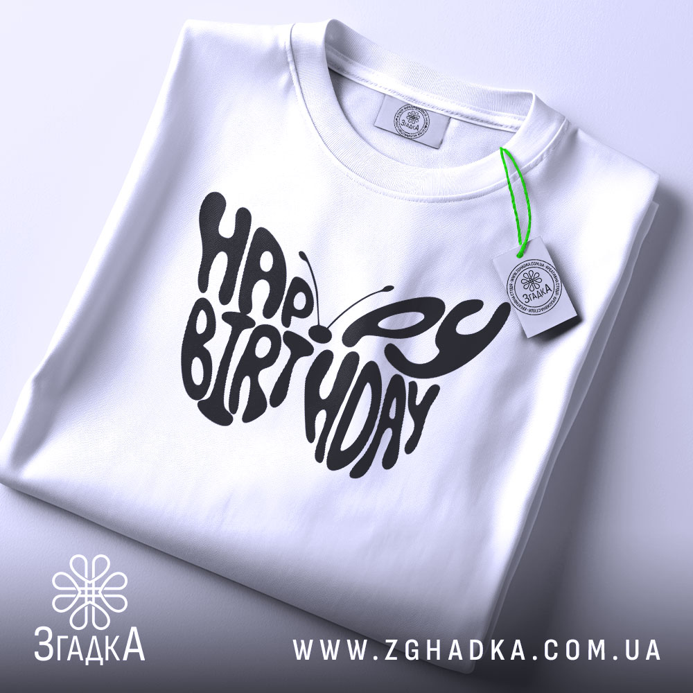 Футболка метелик вітання, біла, складена на поверхні, фон світло-сірий, принт чорного кольору з написом 'HAPPY BIRTHDAY', на футболці є етикетка з зеленим шнурком.
