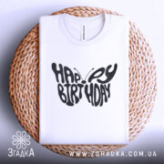 Футболка метелик вітання, біла, складена на плетеній кошику, фон світло-сірий, принт чорного кольору з написом 'HAPPY BIRTHDAY' у формі метелика.