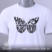 Футболка метелик вітання, біла, складена на поверхні, фон світло-сірий, принт чорного кольору з написом 'HAPPY BIRTHDAY', на футболці є етикетка з зеленим шнурком.