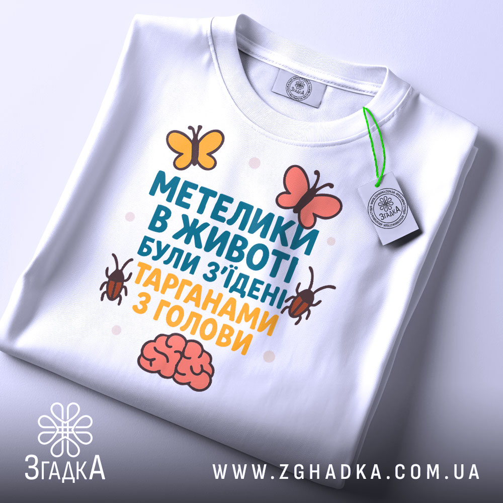 Футболка метелики в животі, білого кольору, складена на поверхні, фон світлий, принт з текстом та ілюстраціями метеликів і мозку, з етикеткою.