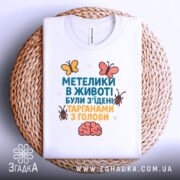 Футболка метелики в животі, білого кольору, складена на кошику з ротанга, фон нейтральний, принт з текстом та малюнками метеликів і мозку.