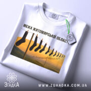 Футболка межа Житомирської області, білого кольору, складена на поверхні, фон світлий, принт з шкарпетками на мотузці з пейзажем, видно етикетку з зеленим шнурком.
