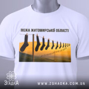 Футболка межа Житомирської області, білого кольору, складена на поверхні, фон світлий, принт з шкарпетками на мотузці з пейзажем, видно етикетку з зеленим шнурком.