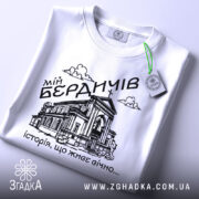 Футболка мій Бердичів, біла, зображена зверху, на плоскій поверхні, фон світло-сірий, принт чорного кольору з зображенням будівлі та написом 'історія, що живе вічно...', з зеленим ярликом.