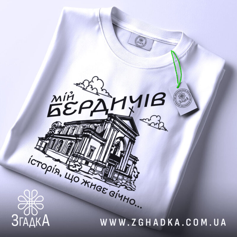 Футболка мій Бердичів, біла, зображена зверху, на плоскій поверхні, фон світло-сірий, принт чорного кольору з зображенням будівлі та написом 'історія, що живе вічно...', з зеленим ярликом.