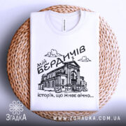 Футболка мій Бердичів, біла, зображена зверху на плетеній кошику, фон світло-сірий, принт чорного кольору з зображенням будівлі та написом 'історія, що живе вічно...'.