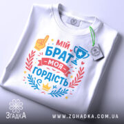 Футболка мій брат моя гордість, біла, складена на поверхні, фон світло-сірий, принт з текстом 'мій брат моя гордість' та ілюстраціями, включаючи жовту руку та кубок, прикрашена червоними та блакитними елементами.