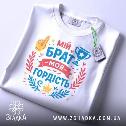 Футболка мій брат моя гордість, біла, складена на поверхні, фон світло-сірий, принт з текстом 'мій брат моя гордість' та ілюстраціями, включаючи жовту руку та кубок, прикрашена червоними та блакитними елементами.