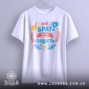 Футболка мій брат моя гордість, біла, підвішена на вішалці, фон світло-сірий, принт з текстом 'мій брат моя гордість' та кольоровими ілюстраціями, включаючи жовту руку, кубок та декоративні елементи.