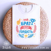 Футболка мій брат моя гордість, біла, складена на плетеній кошику, фон світло-сірий, принт містить текст 'мій брат моя гордість' з жовтою рукою, кубком та декоративними елементами у червоному та блакитному кольорах.