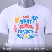 Футболка мій брат моя гордість, біла, складена на поверхні, фон світло-сірий, принт з текстом 'мій брат моя гордість' та ілюстраціями, включаючи жовту руку та кубок, прикрашена червоними та блакитними елементами.