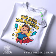 Футболка мій день перемога, біла, складена на столі, фон світло-сірий, принт з хлопчиком у супергеройському костюмі, з тегом на шнурку.