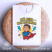 Футболка мій день перемога, біла, складена на плетеному кошику, фон світло-сірий, принт з хлопчиком у костюмі супергероя, оточеним блискавками і написом.