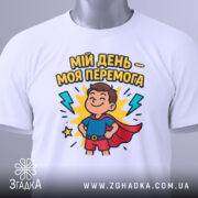 Футболка мій день перемога, біла, складена на столі, фон світло-сірий, принт з хлопчиком у супергеройському костюмі, з тегом на шнурку.