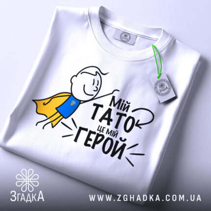 Футболка мій тато герой, біла, зображена зверху, на фоні світло-сірого кольору, принт з малюнком хлопчика в жовтому плащі та текстом "Мій тато це мій герой", на футболці є етикетка з зеленим шнурком.