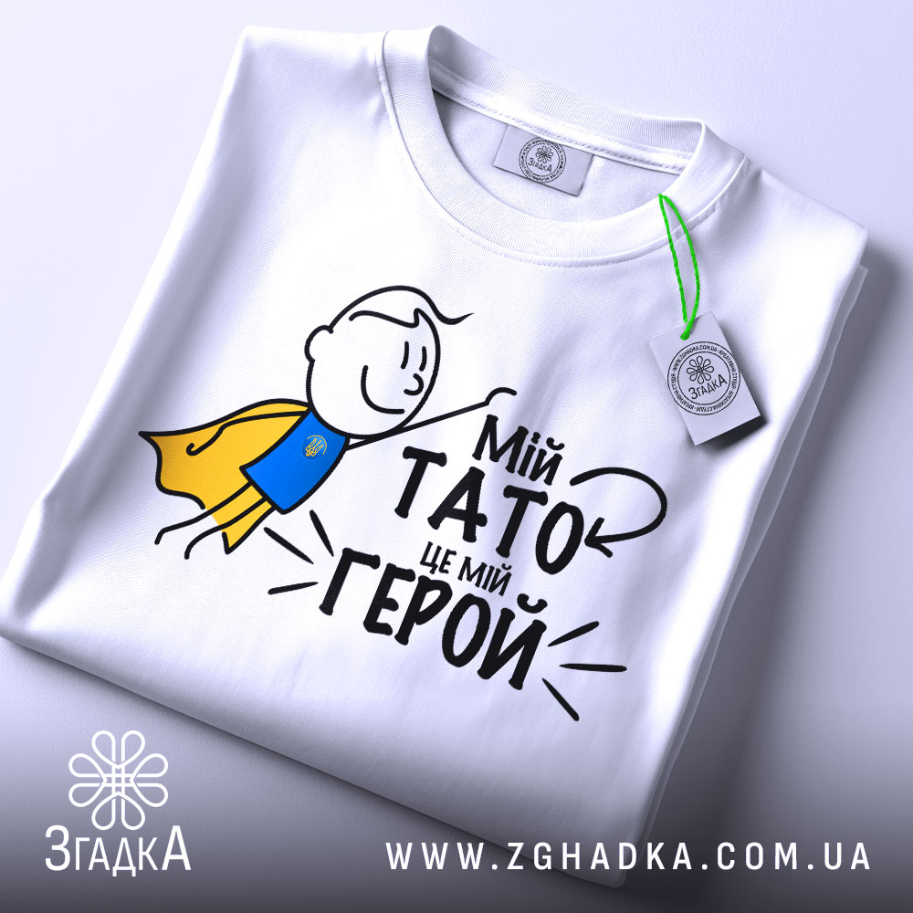 Футболка мій тато герой, біла, зображена зверху, на фоні світло-сірого кольору, принт з малюнком хлопчика в жовтому плащі та текстом "Мій тато це мій герой", на футболці є етикетка з зеленим шнурком.