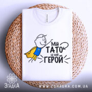 Футболка мій тато герой, біла, зображена зверху на плетеній основі, фон білий, принт з малюнком хлопчика в жовтому плащі та текстом "Мій тато це мій герой".