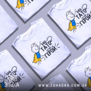 Футболка мій тато герой, біла, зображена з фронтального ракурсу, на фоні світло-сірого кольору, принт з малюнком хлопчика в жовтому плащі та текстом "Мій тато це мій герой".