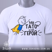Футболка мій тато герой, біла, зображена зверху, на фоні світло-сірого кольору, принт з малюнком хлопчика в жовтому плащі та текстом "Мій тато це мій герой", на футболці є етикетка з зеленим шнурком.