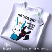 Футболка мій тихий омут. Біла футболка, складена на поверхні, з принтом, що зображує заєць і демон. Принт яскравий, з чіткими кольорами, на фоні світлого кольору. Яскравий етикет з зеленим шнурком.