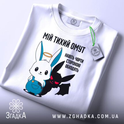 Футболка мій тихий омут. Біла футболка, складена на поверхні, з принтом, що зображує заєць і демон. Принт яскравий, з чіткими кольорами, на фоні світлого кольору. Яскравий етикет з зеленим шнурком.