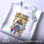 Футболка мій внук білого кольору, складена на поверхні, фон світло-сірий, принт з супергероєм у синій футболці, тримає кубок, з медалями та зірками, на комірці видна етикетка.