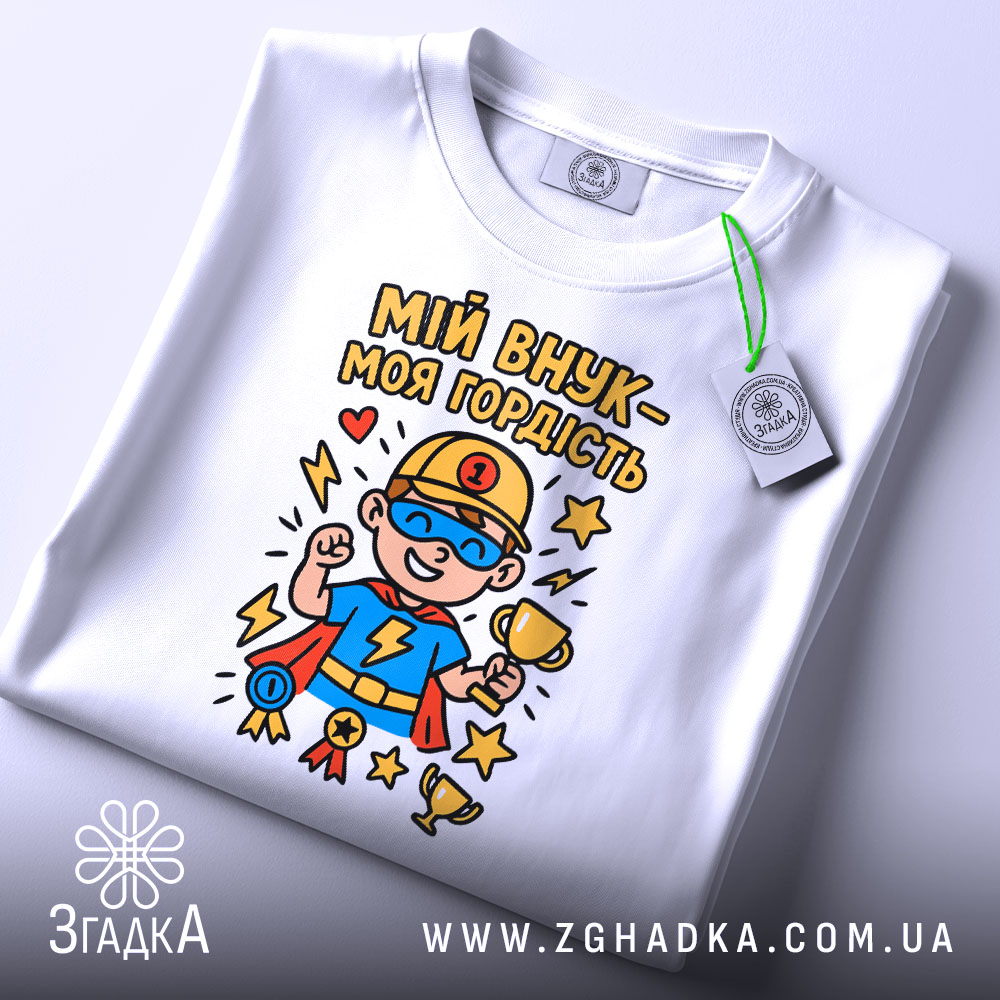 Футболка мій внук білого кольору, складена на поверхні, фон світло-сірий, принт з супергероєм у синій футболці, тримає кубок, з медалями та зірками, на комірці видна етикетка.