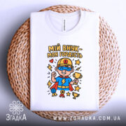 Футболка мій внук білого кольору, складена на плетеній кошику, фон світлий, принт з супергероєм у синій футболці, тримає кубок, оточений зірками, виглядає стильно та привабливо.