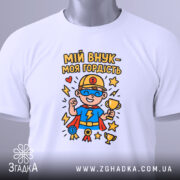 Футболка мій внук білого кольору, складена на поверхні, фон світло-сірий, принт з супергероєм у синій футболці, тримає кубок, з медалями та зірками, на комірці видна етикетка.