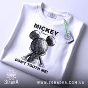 Футболка міккі маус білого кольору, зображена зверху на плоскій поверхні, фон світло-сірий, принт з малюнком міккі мауса ззаду та текстом 'DON'T TOUTH ME!' у чорному кольорі, на футболці є етикетка з зеленим шнурком.