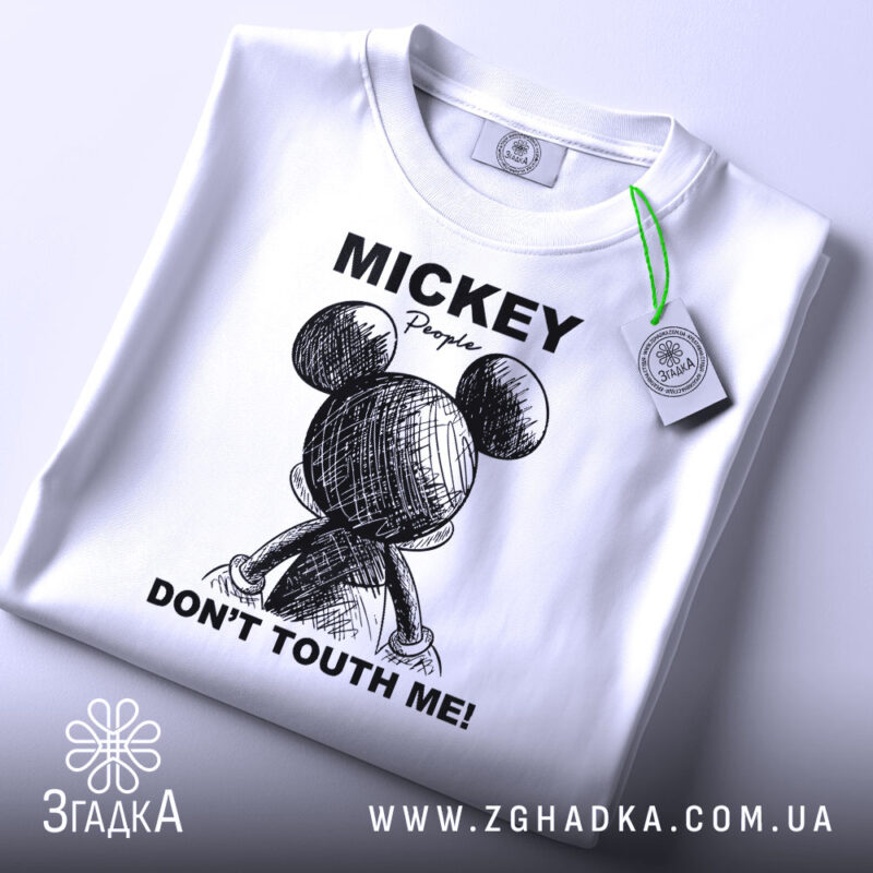 Футболка міккі маус білого кольору, зображена зверху на плоскій поверхні, фон світло-сірий, принт з малюнком міккі мауса ззаду та текстом 'DON'T TOUTH ME!' у чорному кольорі, на футболці є етикетка з зеленим шнурком.