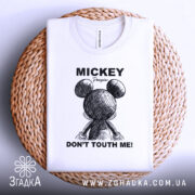Футболка міккі маус білого кольору, зображена зверху на плоскій поверхні з плетеного матеріалу, фон світло-сірий, принт з малюнком міккі мауса ззаду та текстом 'DON'T TOUTH ME!' у чорному кольорі, видно етикетку на горловині.