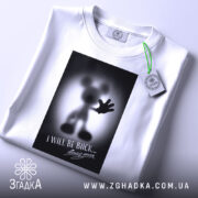 Футболка міккі повертається, біла, складена на столі, фон світло-сірий, принт з силуетом міккі мауса з піднятою рукою та написом "I WILL BE BACK...", на футболці є етикетка з зеленим шнурком.