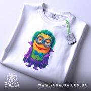 Футболка міньйон злодій джокер, біла, складена на столі, фон світлий, принт з міньйоном у фіолетовому пальто, з зеленим волоссям та окулярами, з етикеткою з зеленим шнурком.