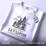 Футболка місто Бердичів подарунок, біла, складена на поверхні, фон світло-сірий, принт з архітектурним зображенням церкви, з зеленим ярликом.