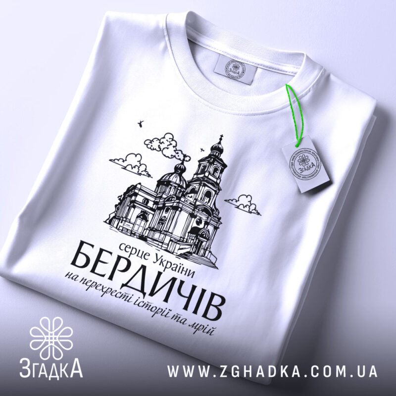 Футболка місто Бердичів подарунок, біла, складена на поверхні, фон світло-сірий, принт з архітектурним зображенням церкви, з зеленим ярликом.