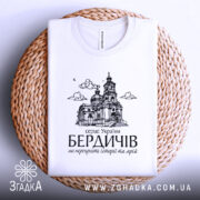 Футболка місто Бердичів подарунок, біла, складена на плетеній кошику, фон білий, принт з архітектурним зображенням церкви та текстом на українській мові.