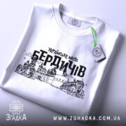 Футболка місто Бердичів, біла, зображена зверху, складена на поверхні, фон світлий, принт містить чорний контурний малюнок архітектурних пам'яток Бердичева з написом 'УКРАЇНСЬКЕ МІСТО БЕРДИЧІВ', на футболці є етикетка з зеленим шнурком.